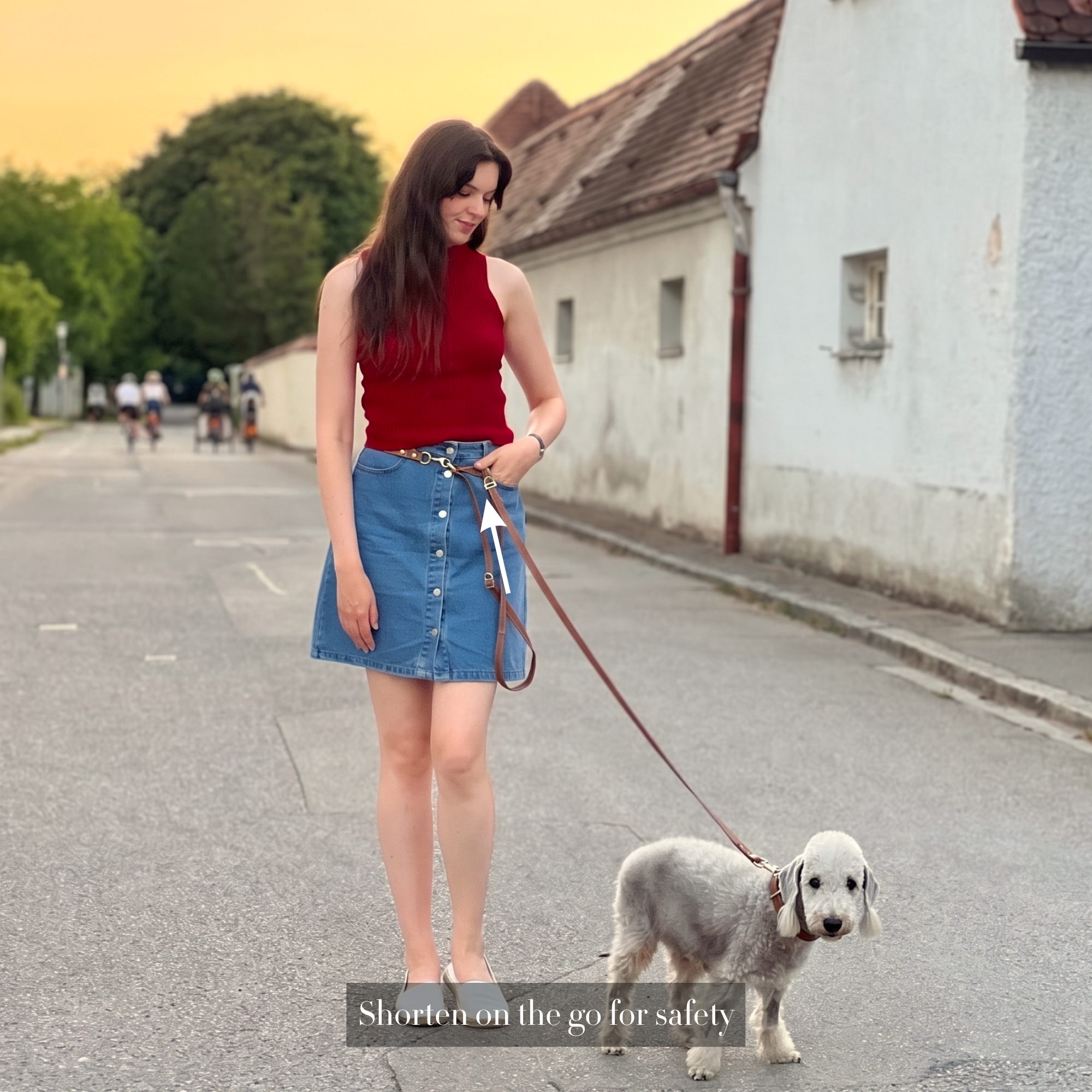 Woman in red top and blue skirt walking a dog on a handsfree leather leash in a village setting. #lang_en