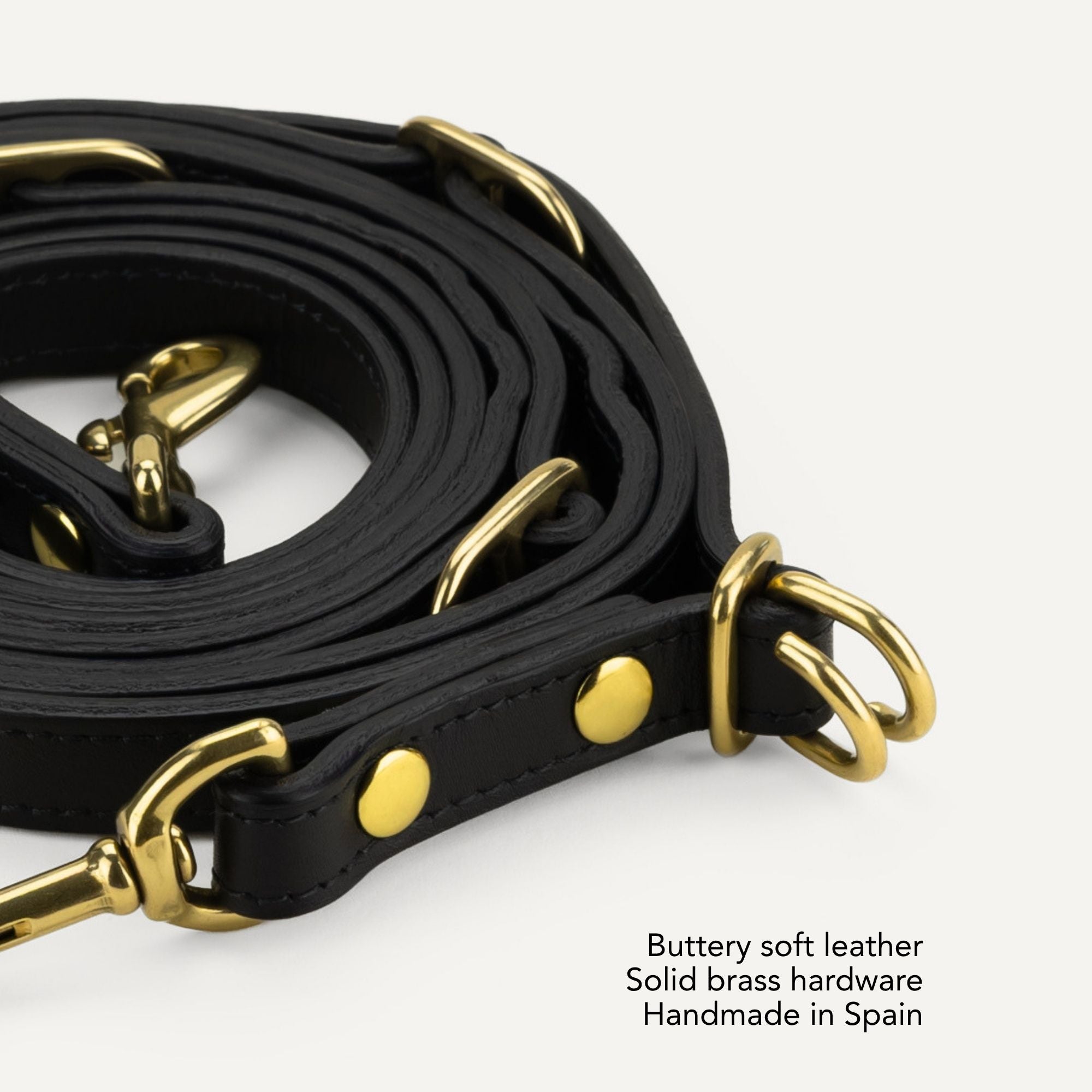 A close-up of a black leather strap with polished brass hardware, coiled on a light gray background with text noting 'Buttery soft leather', 'Solid brass hardware', and 'Handmade in Spain'. #lang_en