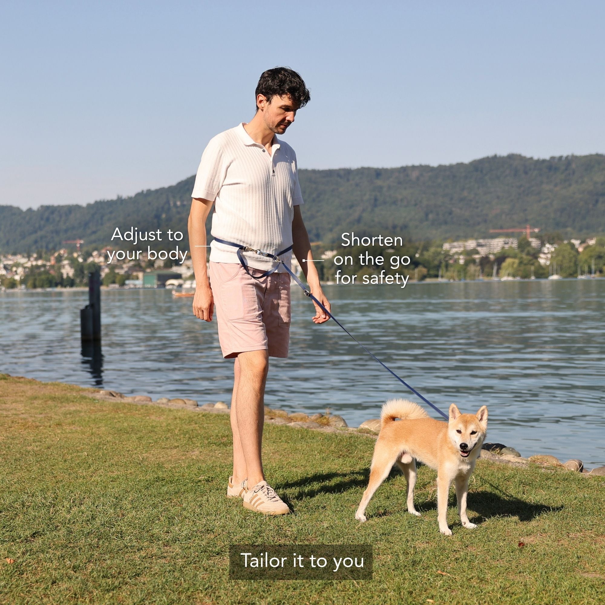 A man walks a Shiba Inu dog on a grassy lakeside with overlaid text explaining how to tailor the dog leash: 'Adjust to your body', 'Shorten on the go for safety', and 'Tailor it to you'. #lang_en