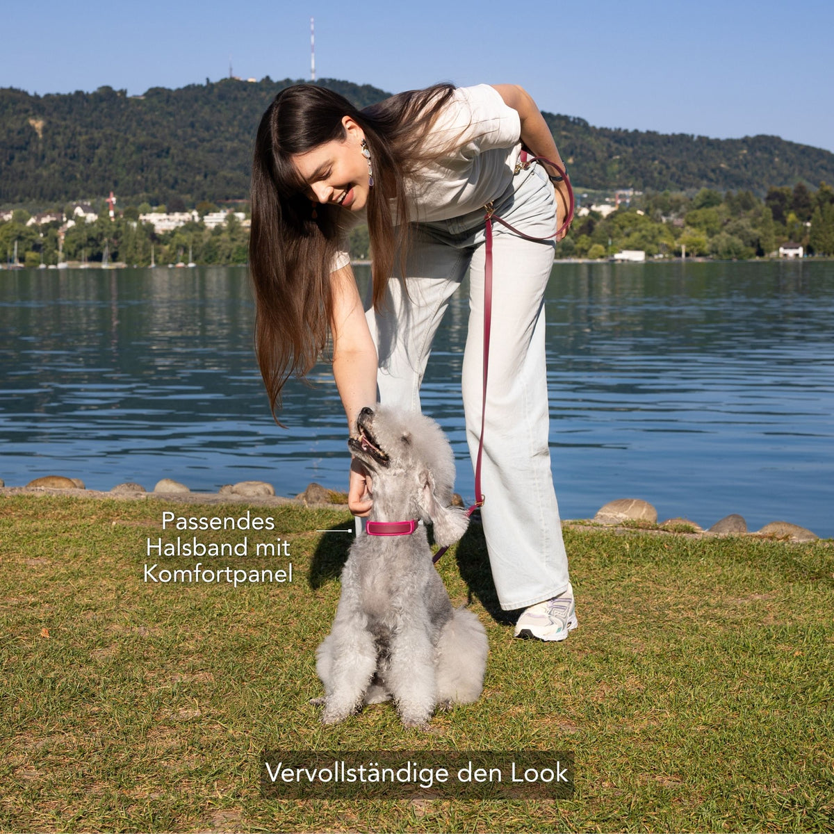 A woman outdoors by a lake bends down to interact with a small gray dog wearing a pink collar and leash. Text on the image highlights the matching collar with a comfort panel and says to complete the look. #lang_de