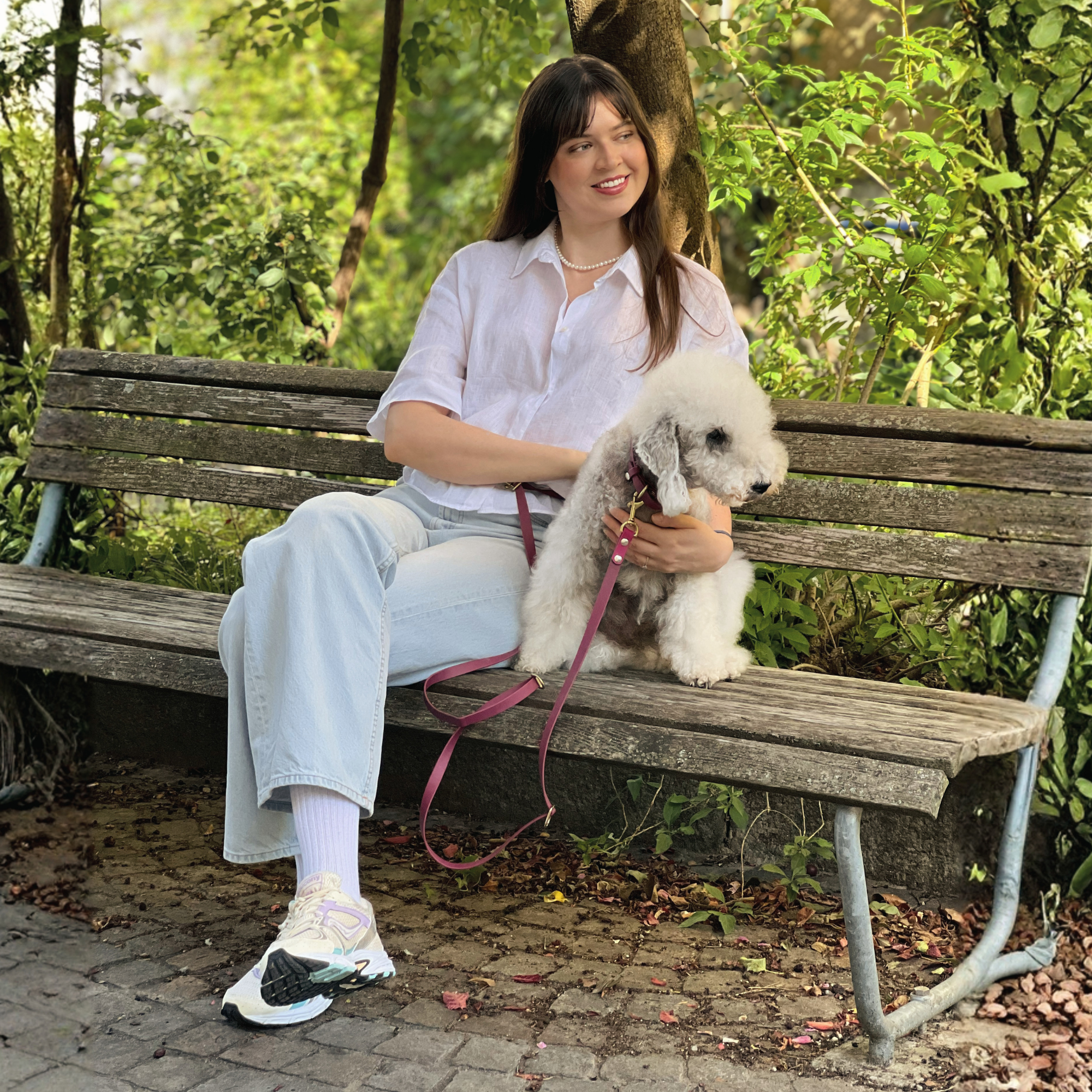 Woman sitting on a bench with a small white dog in a park with a around-the-waist handsfree dog leash and matching collar from BioThane in Pink.