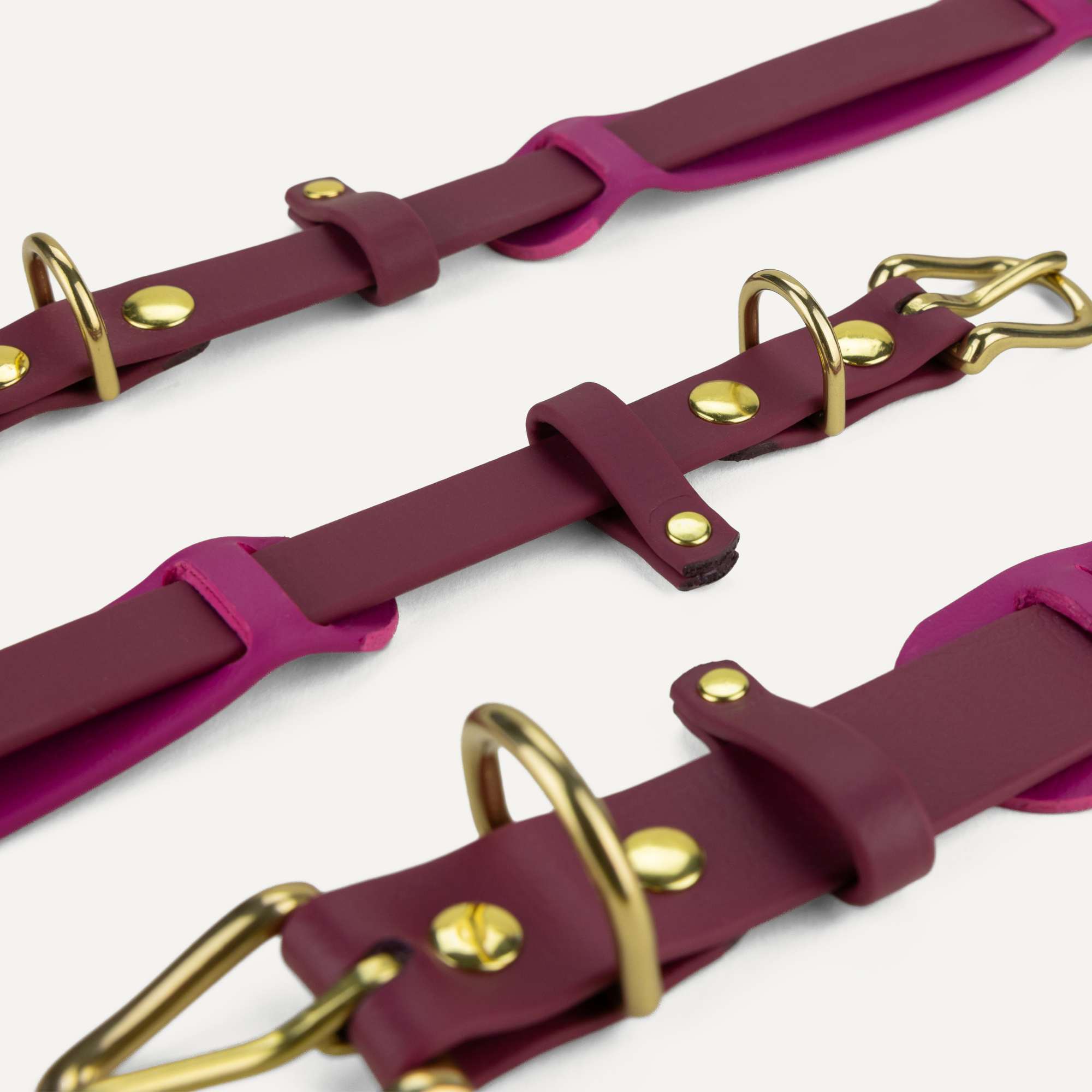 A close-up of three matching burgundy straps with gold-tone hardware, including metal rings and rivets, arranged diagonally on a light off-white background.