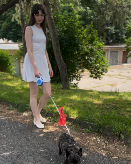 Woman in a white dress walking a small black dog on a leash in a park.
