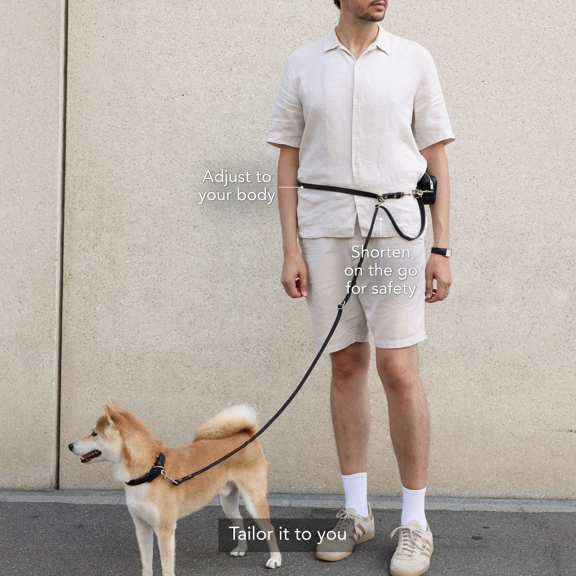 A man wearing a beige short-sleeve shirt and shorts stands outdoors next to a Shiba Inu dog, demonstrating a black waist leash belt. The leash is attached to the belt and extends to the dog's collar. #lang_en
