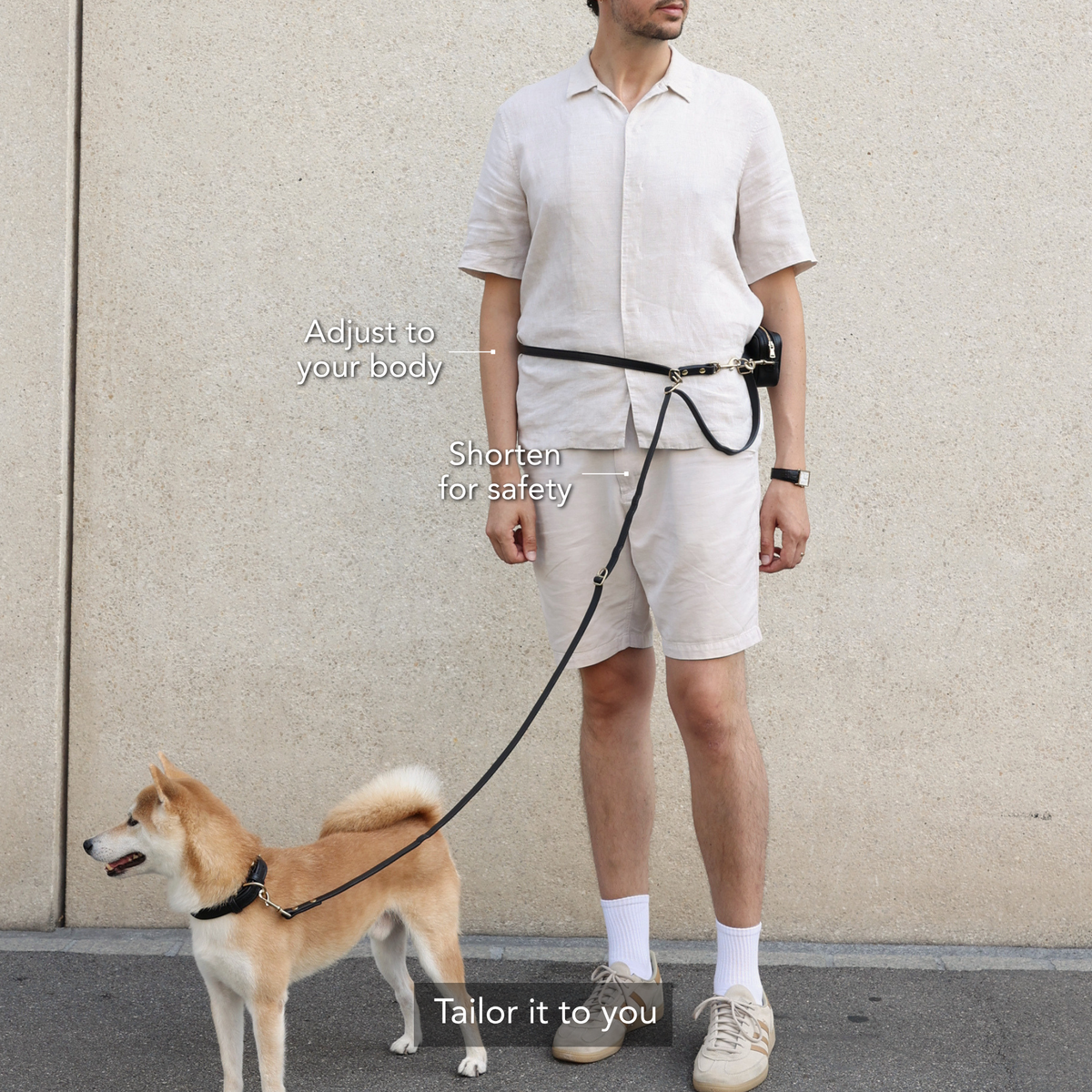 A man wearing a beige short-sleeve shirt and shorts stands outdoors next to a Shiba Inu dog, demonstrating a black waist leash belt. The leash is attached to the belt and extends to the dog's collar. #lang_en