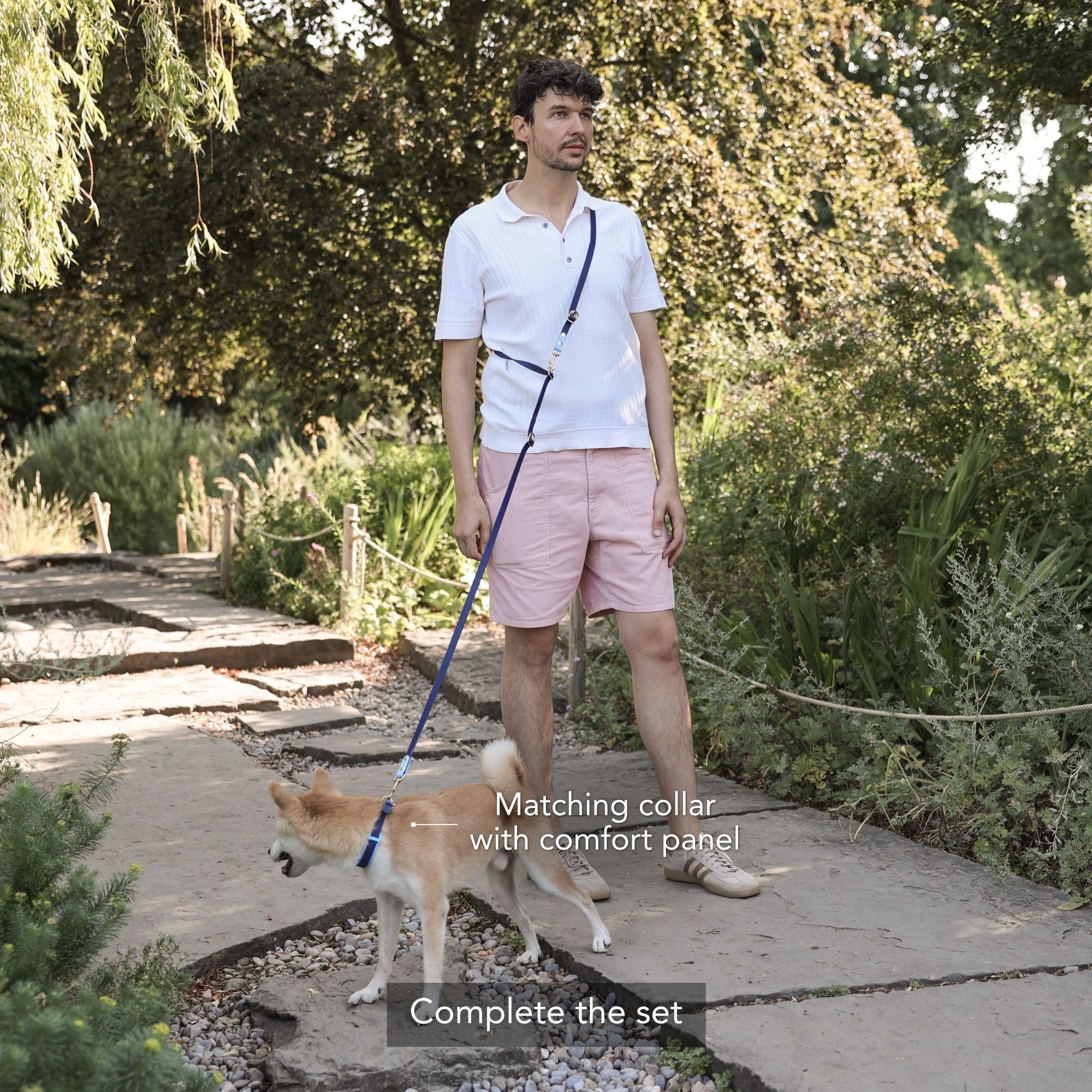 A man wearing a white polo shirt and pink shorts stands on a stone path in a garden, holding a blue dog leash with a matching blue collar worn by a small tan dog. Text overlays read 'Matching collar with comfort panel' and 'Complete the set'. #lang_en