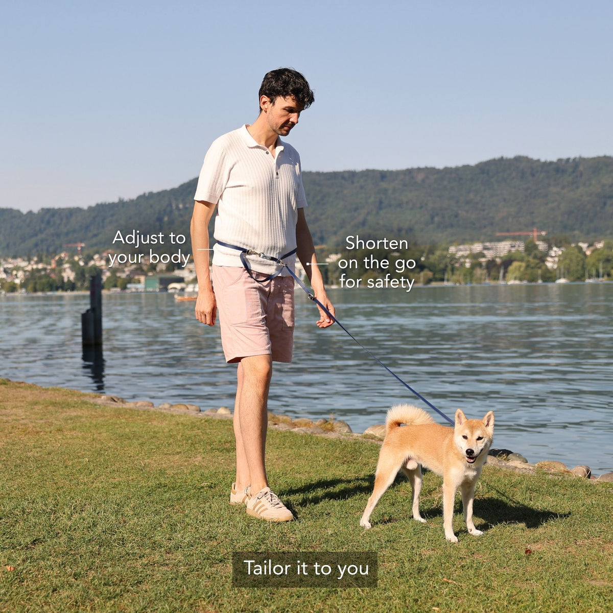 A man walks a Shiba Inu dog on a grassy lakeside with overlaid text explaining how to tailor the dog leash: 'Adjust to your body', 'Shorten on the go for safety', and 'Tailor it to you'. #lang_en