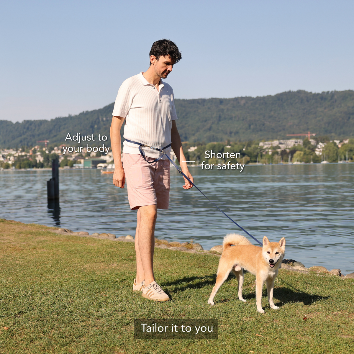 A man walks a Shiba Inu dog on a grassy lakeside with overlaid text explaining how to tailor the dog leash: 'Adjust to your body', 'Shorten on the go for safety', and 'Tailor it to you'. #lang_en