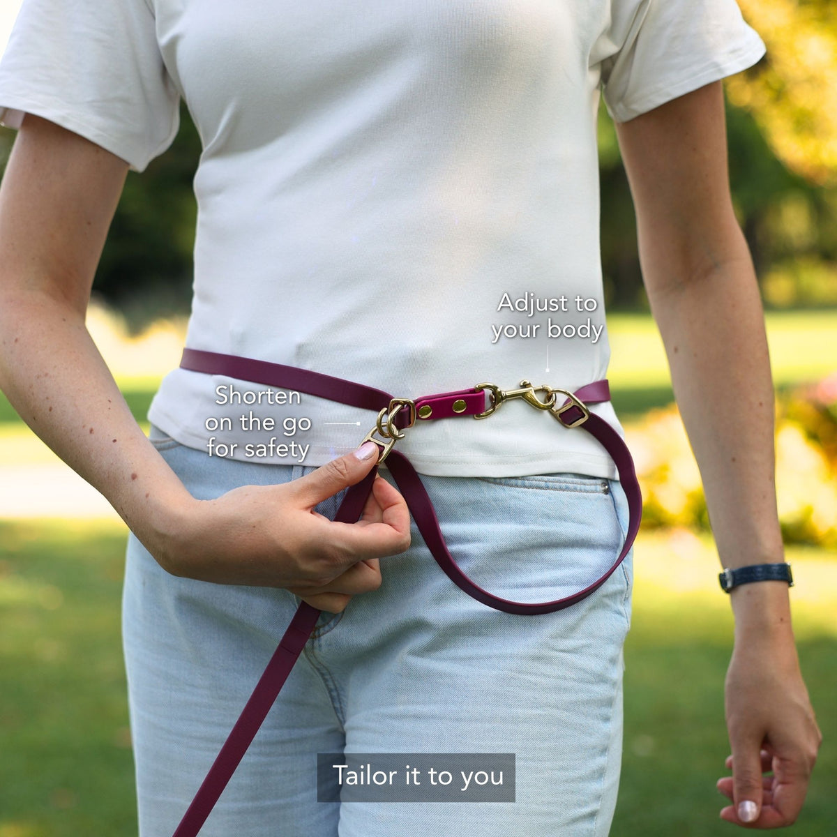 A person outdoors wearing a burgundy leather belt-style pet leash with gold-tone hardware, shown being adjusted around the waist. White text callouts read 'Shorten on the go for safety', 'Adjust to your body', and 'Tailor it to you'. #lang_en