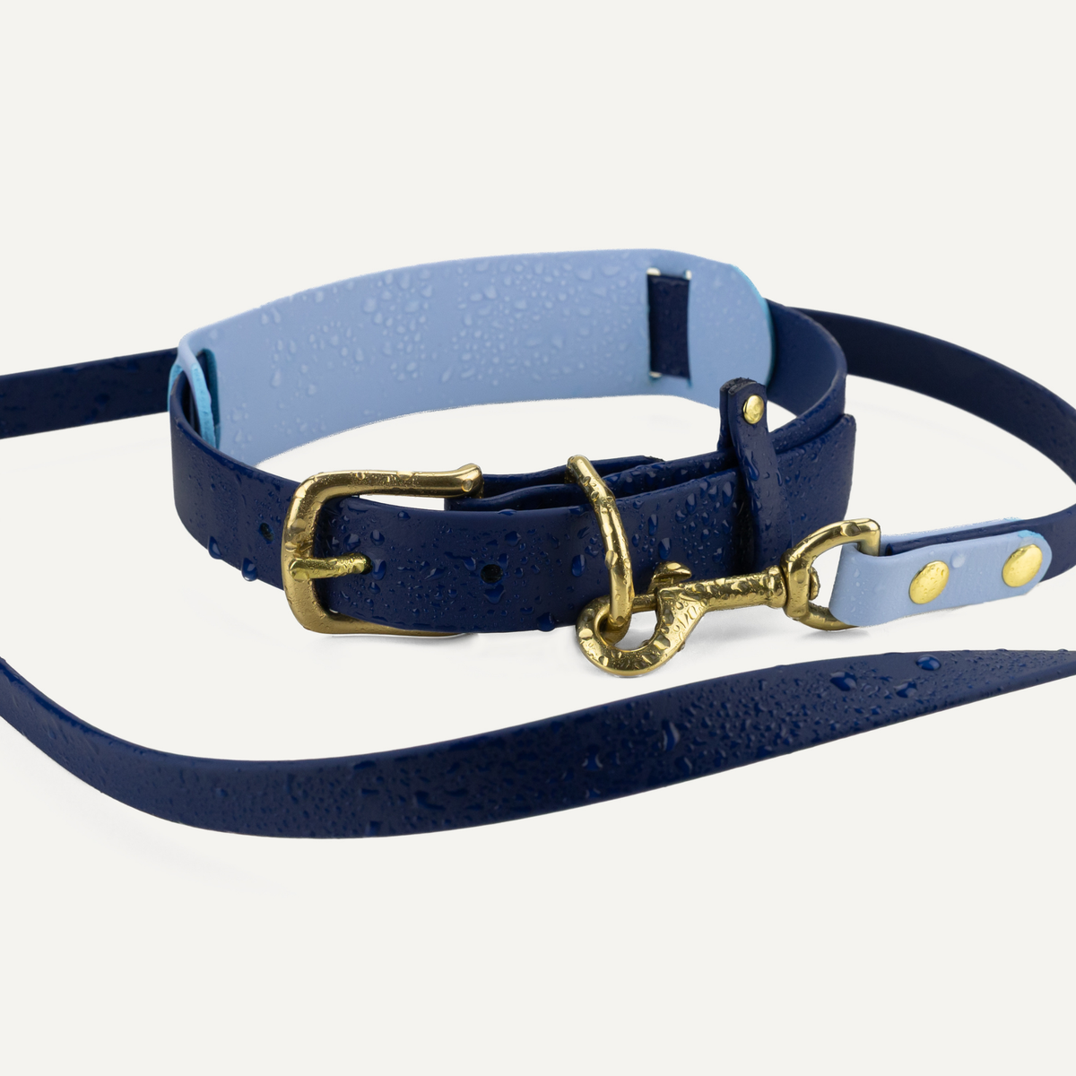 A navy blue leather dog collar with a matching long lead strap, featuring a light blue underside and polished gold-tone metal hardware, shown on a light off-white background with water droplets.