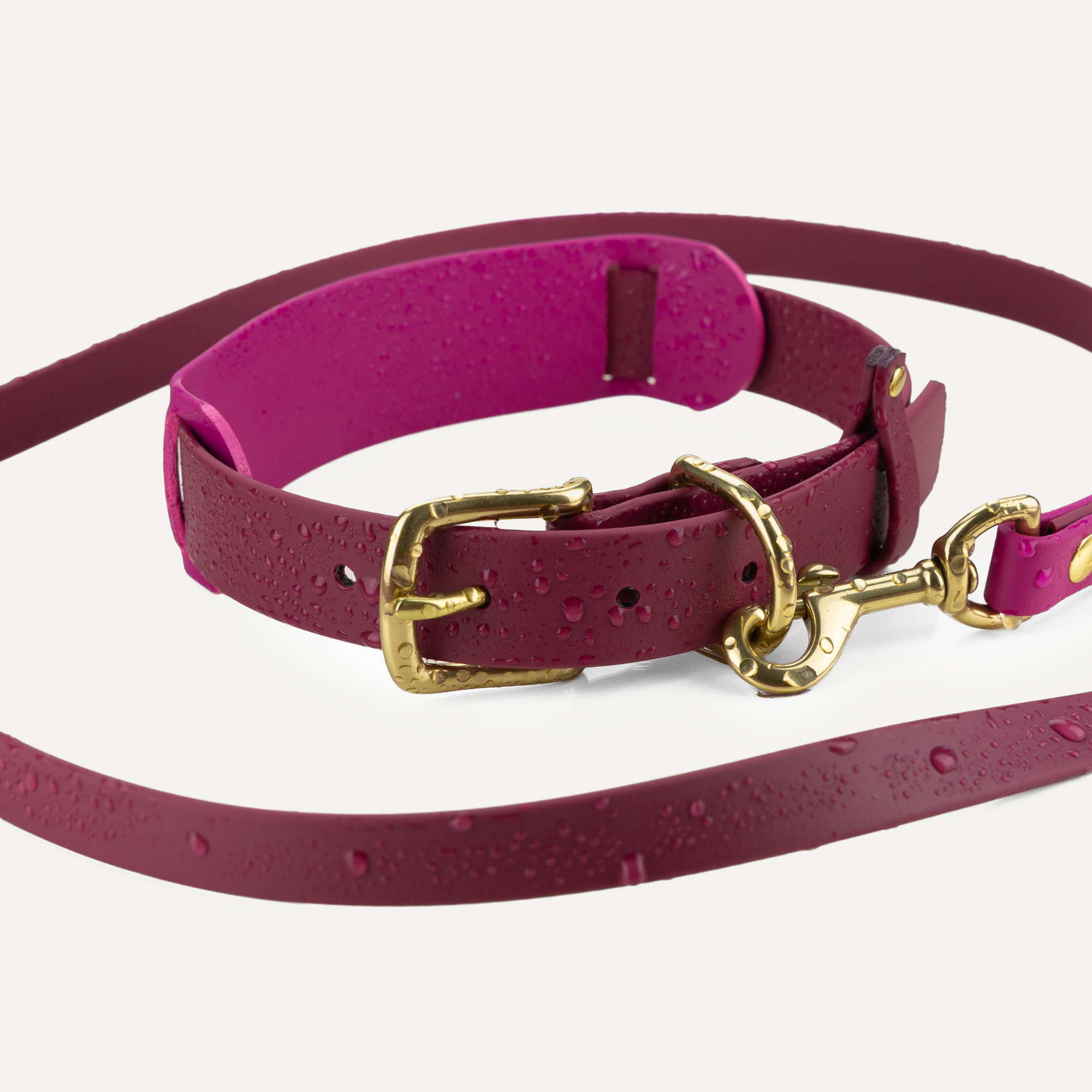A magenta pink biothane dog collar and matching leash with gold-tone hardware, shown on a light off-white background with water droplets on the surface.