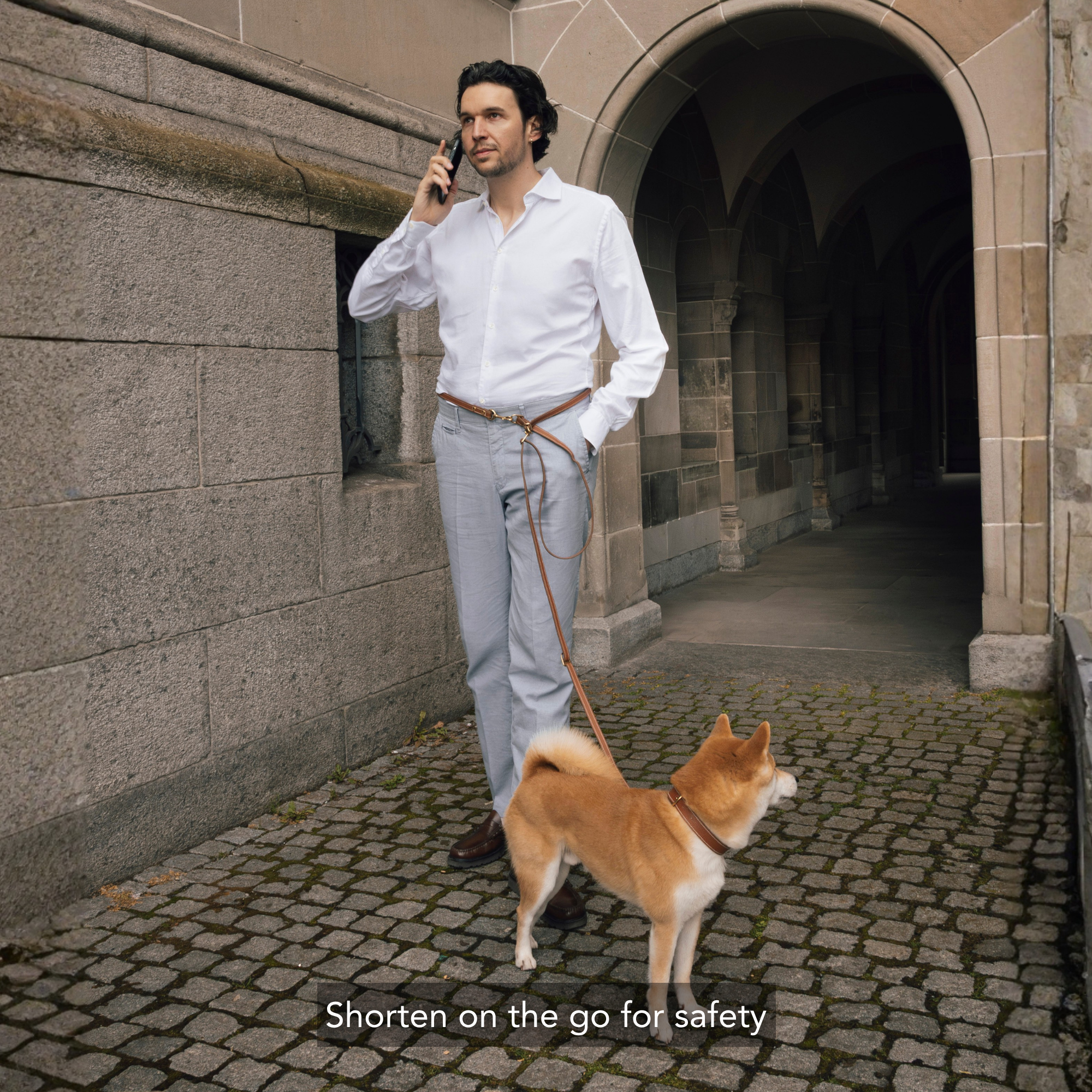 A man in a white dress shirt and light gray trousers stands on a cobblestone walkway next to a stone building, talking on a mobile phone while wearing a tan handsfree leash attached to a Shiba Inu dog. White text at the bottom reads 'Shorten on the go for safety'. #lang_en